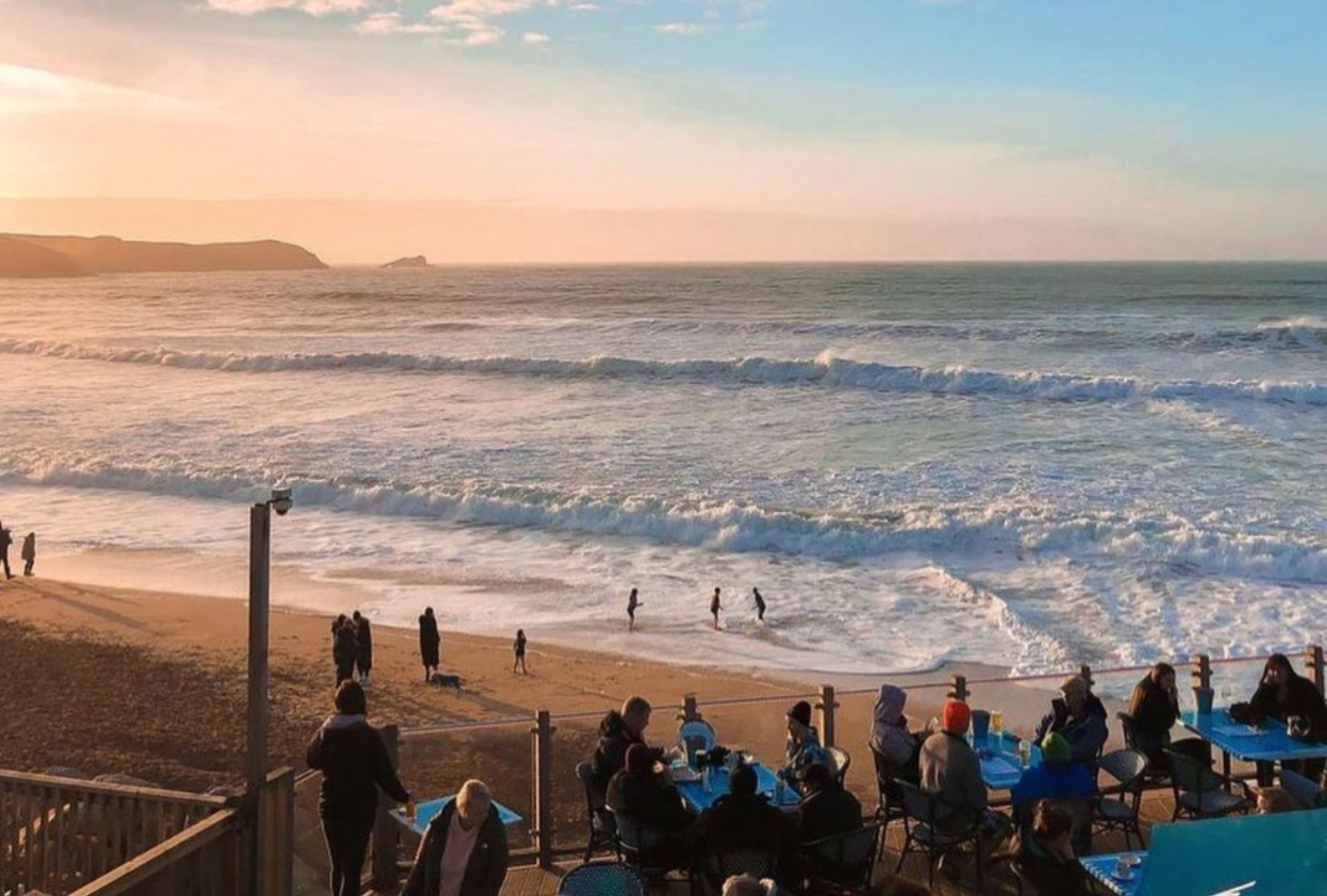 The sunset on fistral beach from the point of view of fistral bear bar in newquay. people sit on tables enjoying a drink and children play in the surf