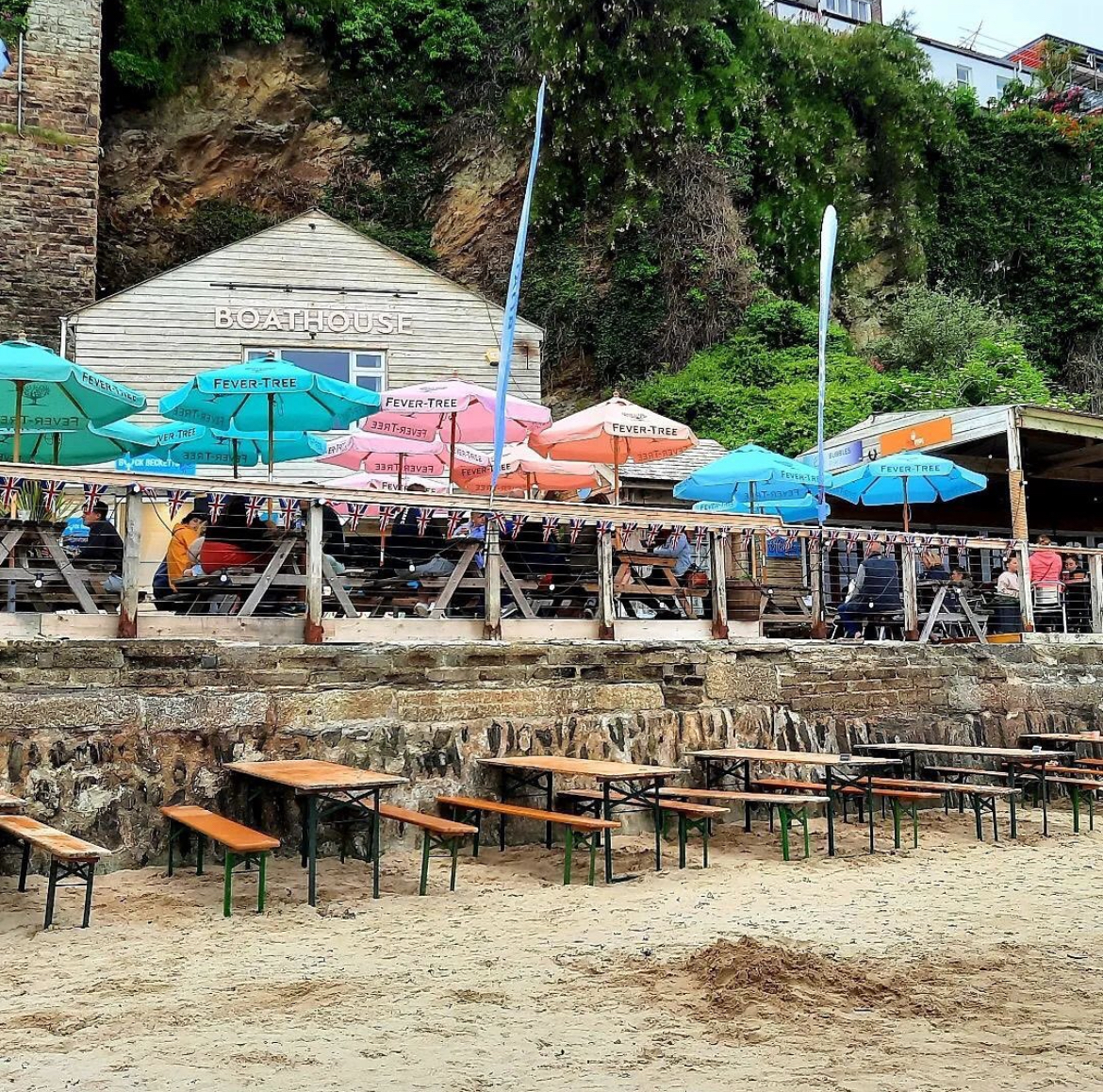 A view from the harbour across the sand to the boathouse bar with colourful umbrellas and outside seating