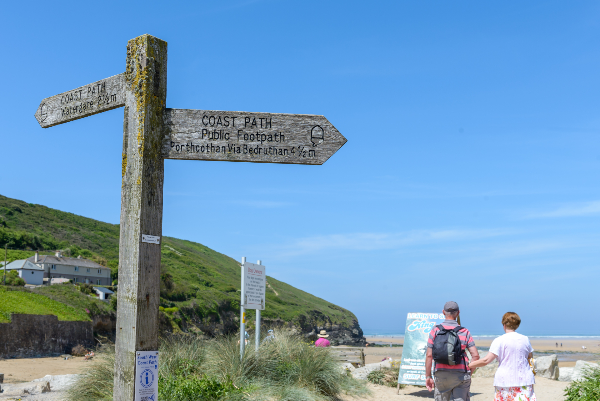 A couple walk down to mawgan porth beach past the south west coast path sign on a sunny day