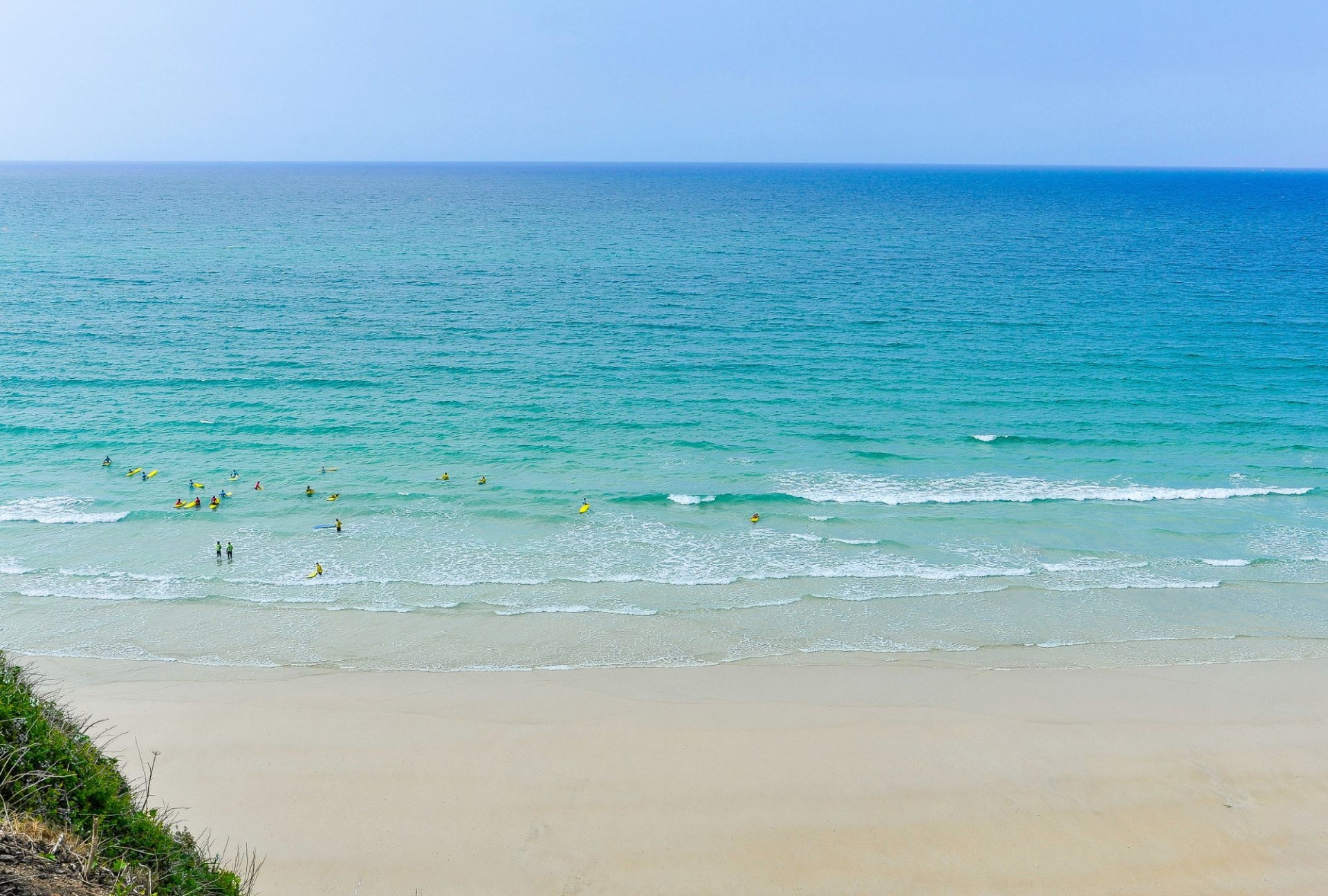 A sunny day on great western beach in Newquay, Cornwall. There's golden sand, turquoise ocean and some surfers dotted about in the water waiting to catch a a wave. You can see a small piece of cliff edge in the bottom left-hand corner