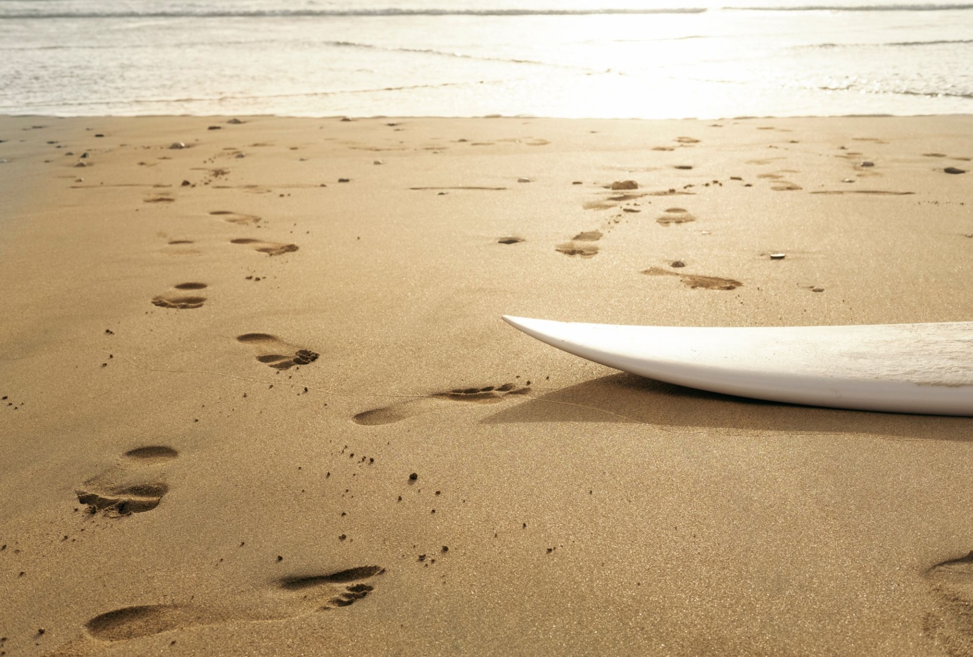 The angled head of a white surfboard sits on the right hand side of this shot. The surfboard is laying on the sand on Fistral beach in Newquay, an there are several footprints in the sand. The sun glows brightly over the sea in the background, and it seems like it will be sunset soon.