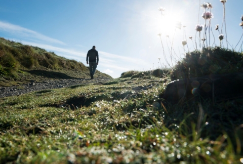 A man walks the south west coast path in Cornwall on a sunny spring day, with grass and flowers in the foreground and a silhouette of a walker on the left hand side walking away from the camera